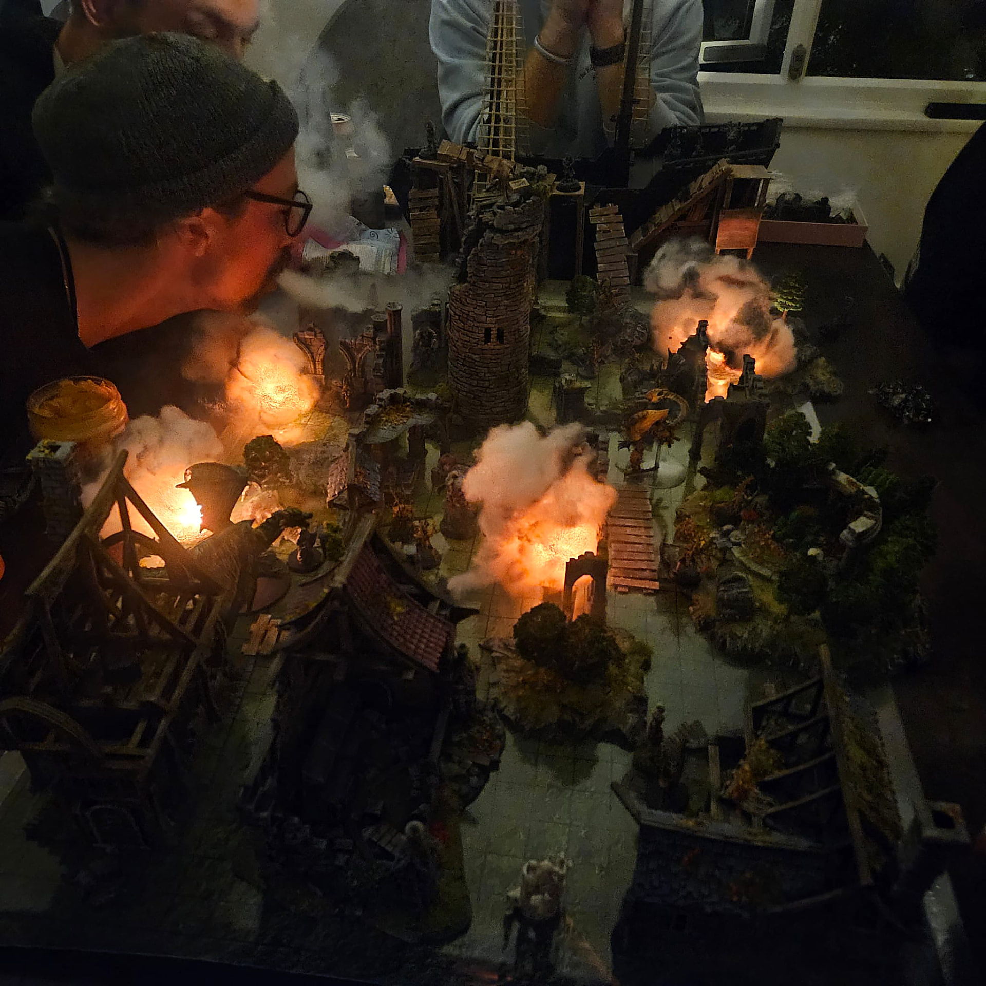 A tabletop village lit by flickering fire effects and drifting cotton smoke